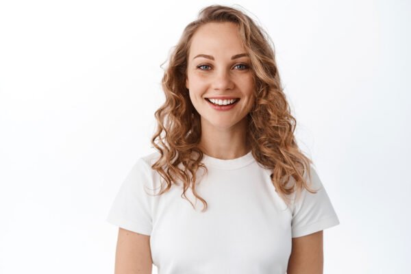 Smiling blond woman with white perfect smile and natural face, looking happy and confident at camera, standing in t-shirt against white background.